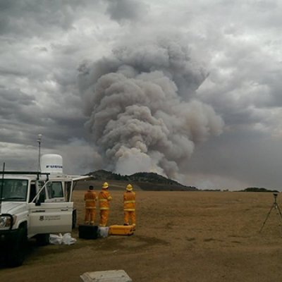 Firefighters standing near a truck watching a large plume of smoke.